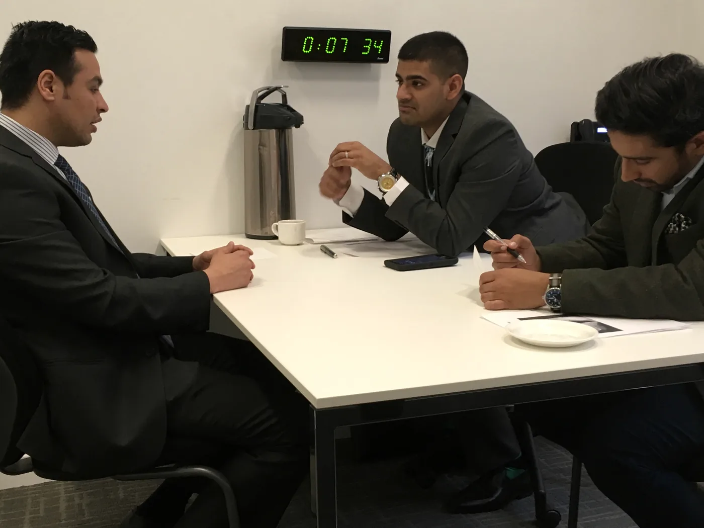 A candidate answering two examiners during a mock interview, with the station clock on the wall